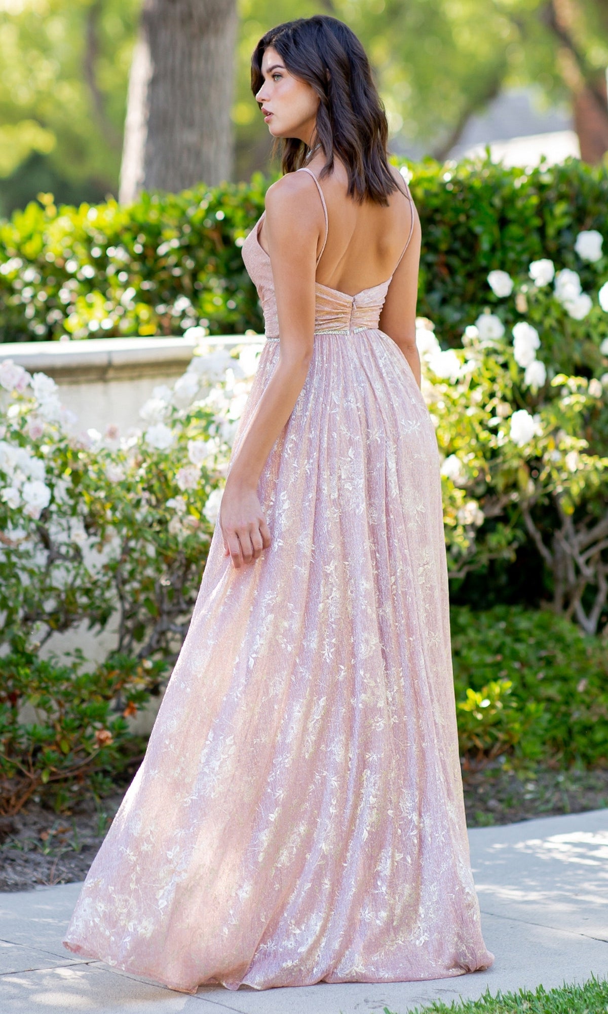 A woman wearing the Cinderella Couture 8086J Blush Pink Prom Dress stands outdoors on a walkway, surrounded by green bushes and white flowers.